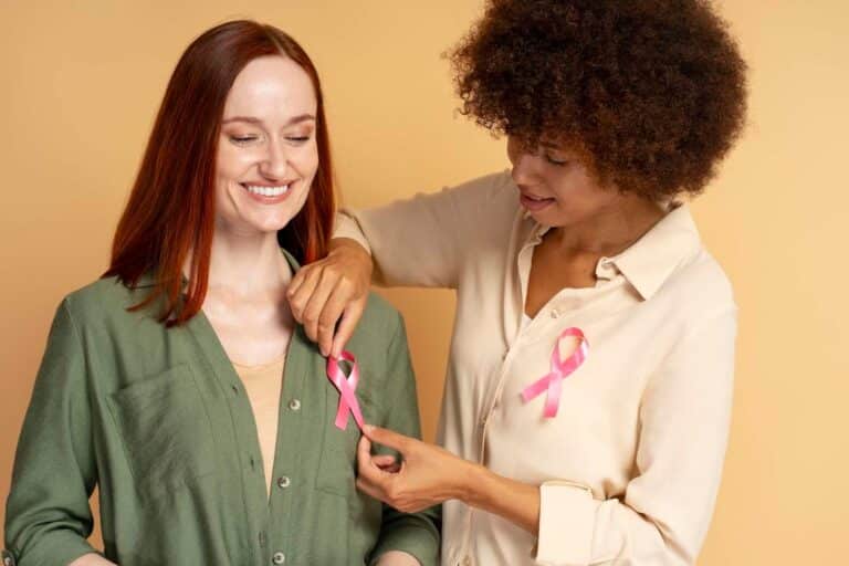 Two women smiling while pinning pink ribbons on their shirts for breast cancer awareness